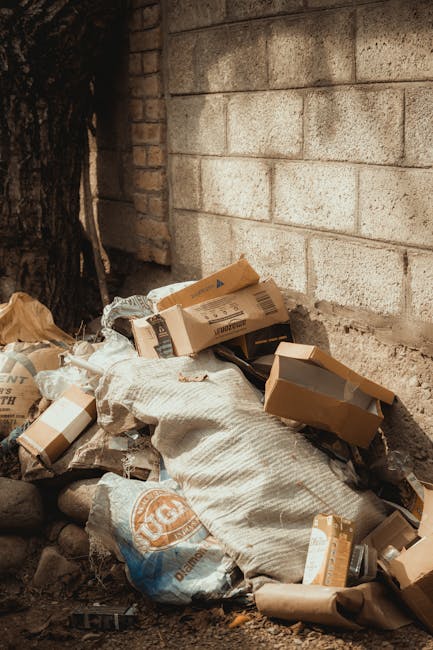 A narrow urban alleyway filled with accumulating rubbish and discarded items. In the foreground, a large, weathered roll-top refuse bag with visible creases and dirt stains stands upright on the uneve