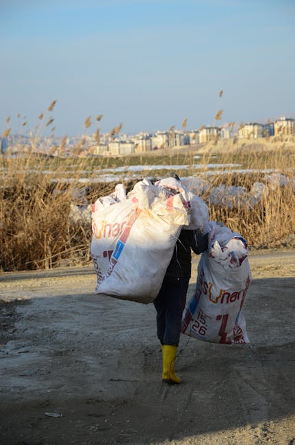 A person dressed in dark clothing and wearing yellow rubber boots is walking along a dirt path carrying large white plastic rubbish bags filled with waste, with some bags showing partial red and blue 